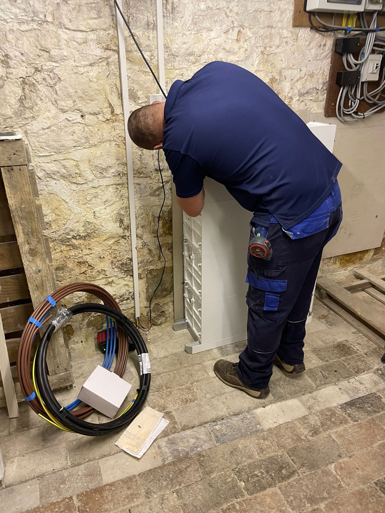 Electrician in blue uniform working on electrical panel in a basement. Cables and tools on the floor.
