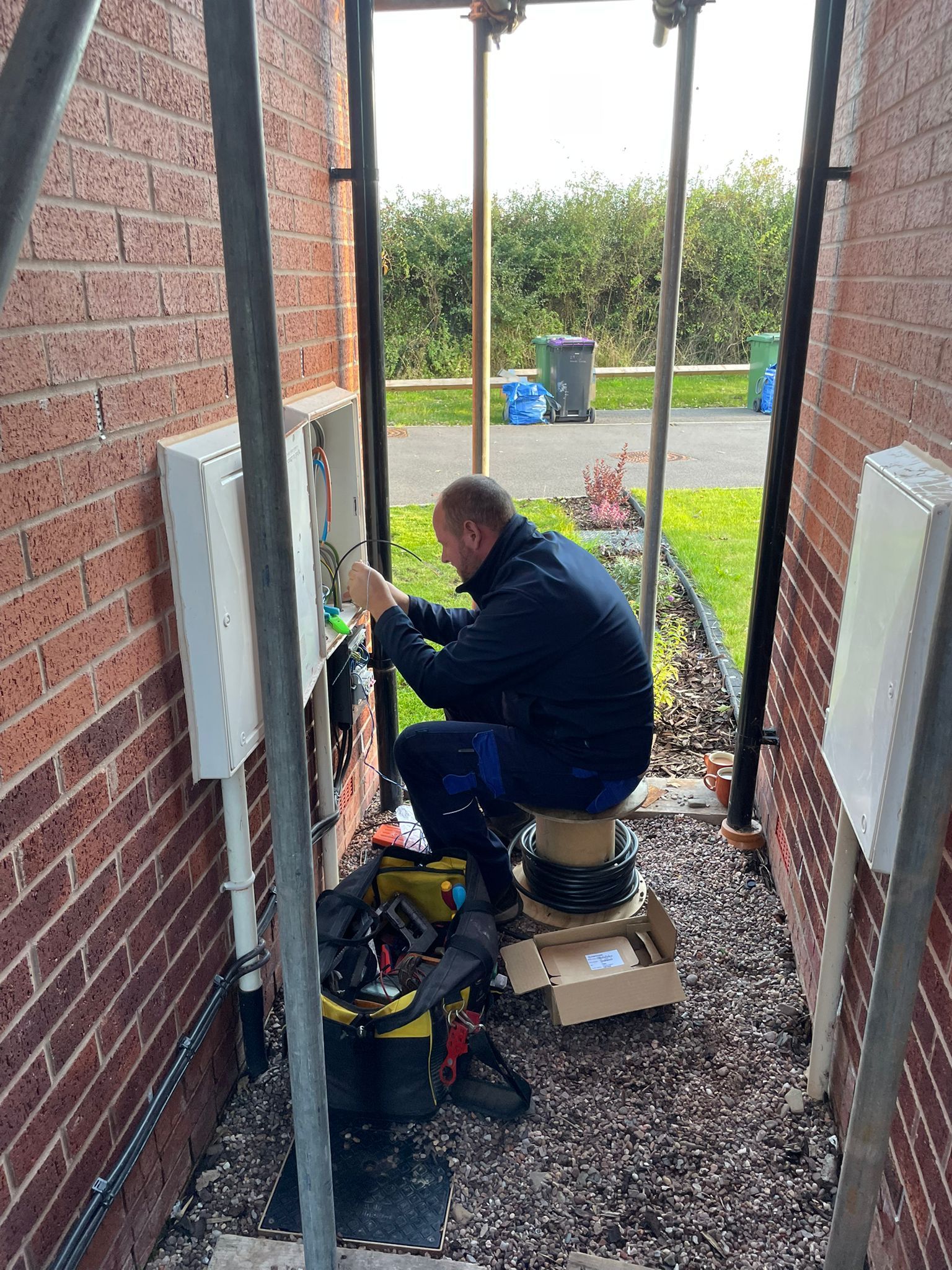Man wiring electrical boxes, working in a narrow outdoor space between brick walls.