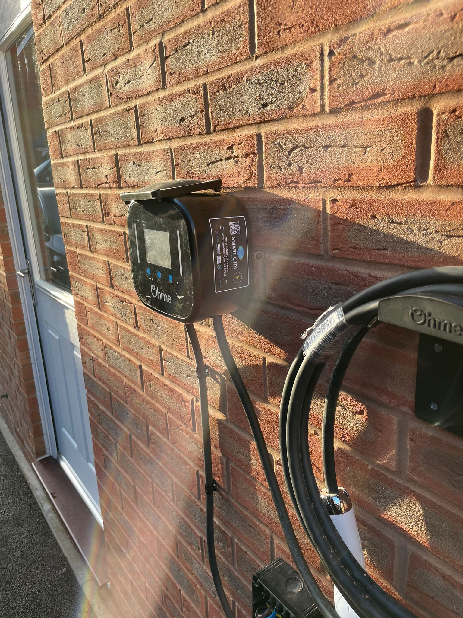 Electric vehicle charger mounted on a red brick wall next to a white door. A charging cable is connected.