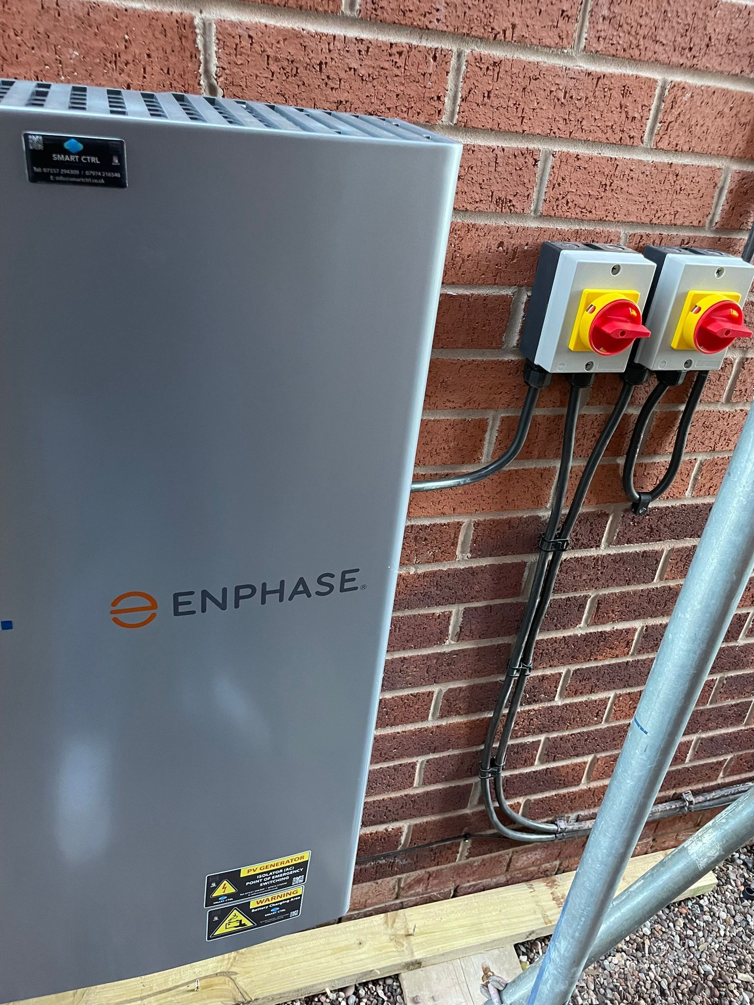 Enphase solar battery unit on a brick wall with two red-handled electrical disconnects.