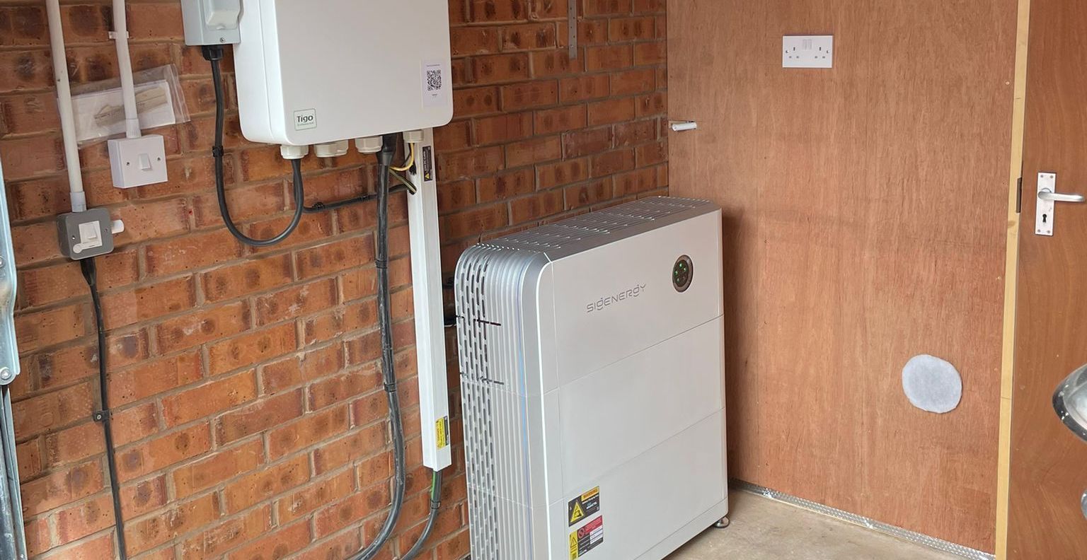 Solar panel equipment, including an inverter and a battery, installed indoors against a brick wall and a plywood wall.