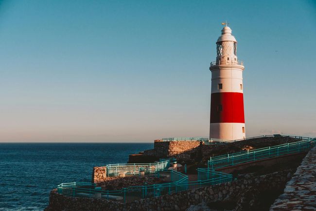 White and red lighthouse on a rocky coast under a clear blue sky, overlooking the sea.