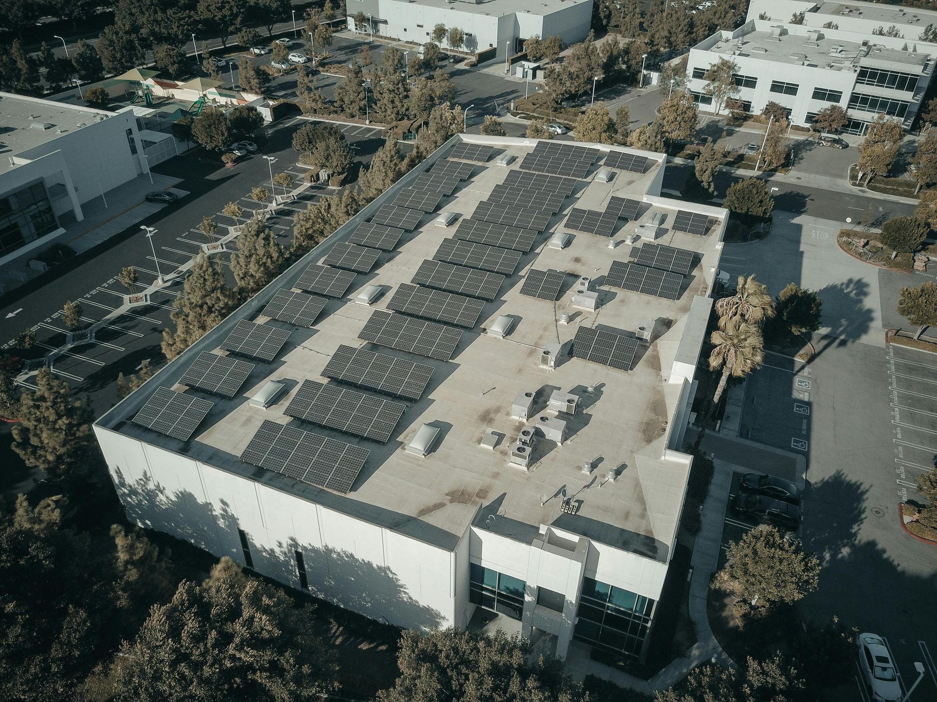 Overhead view of a building with a flat roof covered in solar panels, surrounded by trees and other buildings.