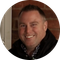 Smiling man with short dark hair, wearing a dark jacket over a plaid shirt, standing in front of a brick wall.