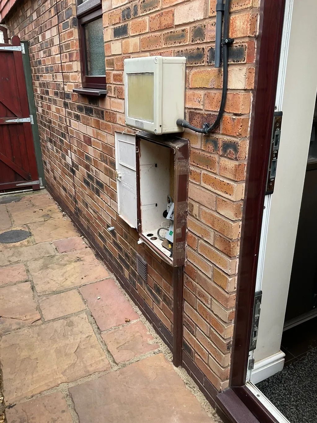 Brick building exterior with open utility panel and white box above, narrow alley.