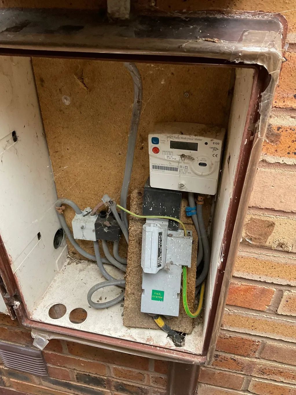 Electric meter and fuse box within an outdoor enclosure, mounted on a brick wall.