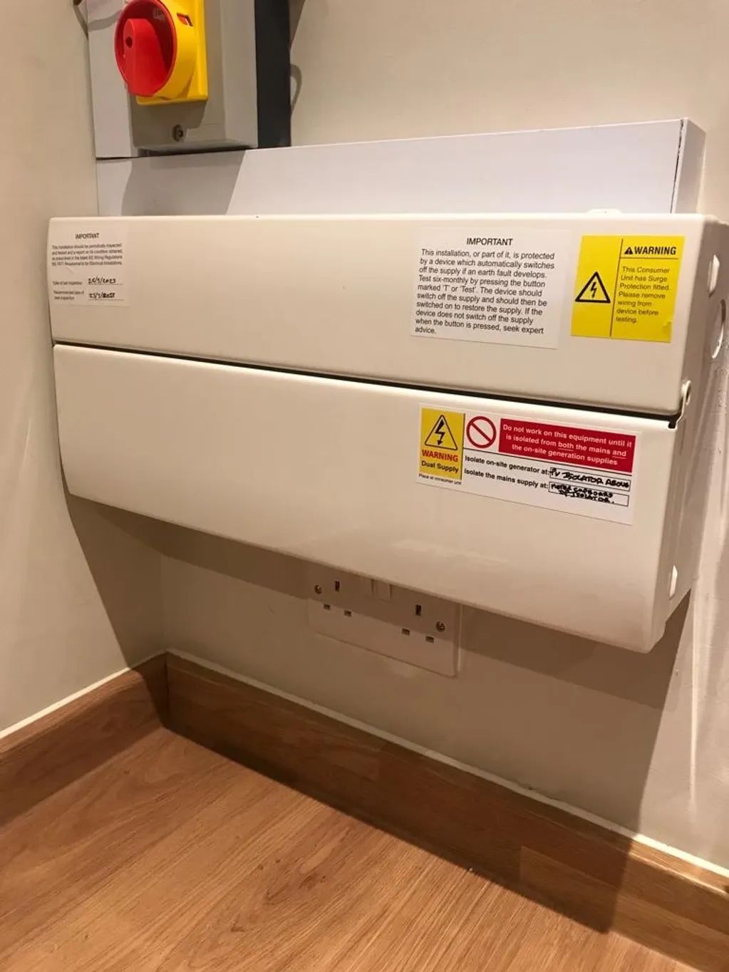 White, wall-mounted, fold-down changing table with safety signs and electrical outlet. Wooden floor and wall.