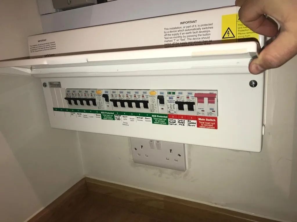 Hand opening a white electrical fuse box on a beige wall, revealing switches and a double outlet.