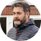Man with a beard in a blue jacket, standing outdoors in front of a brick building.