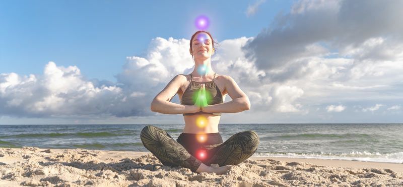 Woman meditating on a beach with colored chakras over body, sunny sky, and ocean. Understaing Energy Healing An introduction to the Health and Wellness Benefits of Energy Healing