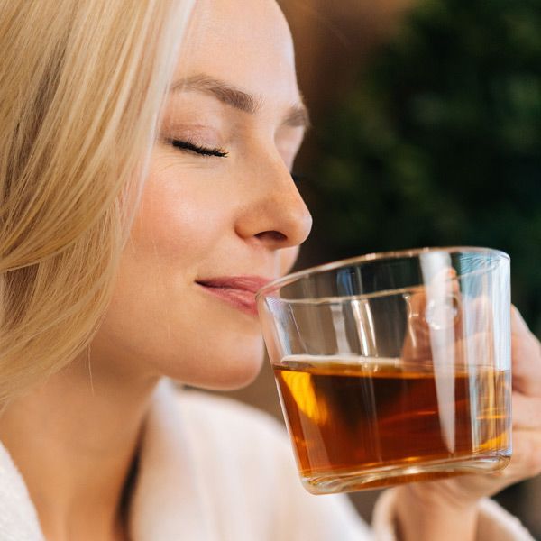 Woman enjoying the aroma of tea in a clear glass. She has blonde hair and her eyes are closed.