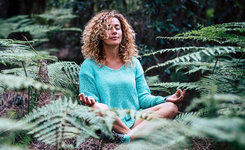 A woman is sitting in a lotus position in the woods surrounded by ferns Grounding with Nature for Healing and Well-Being