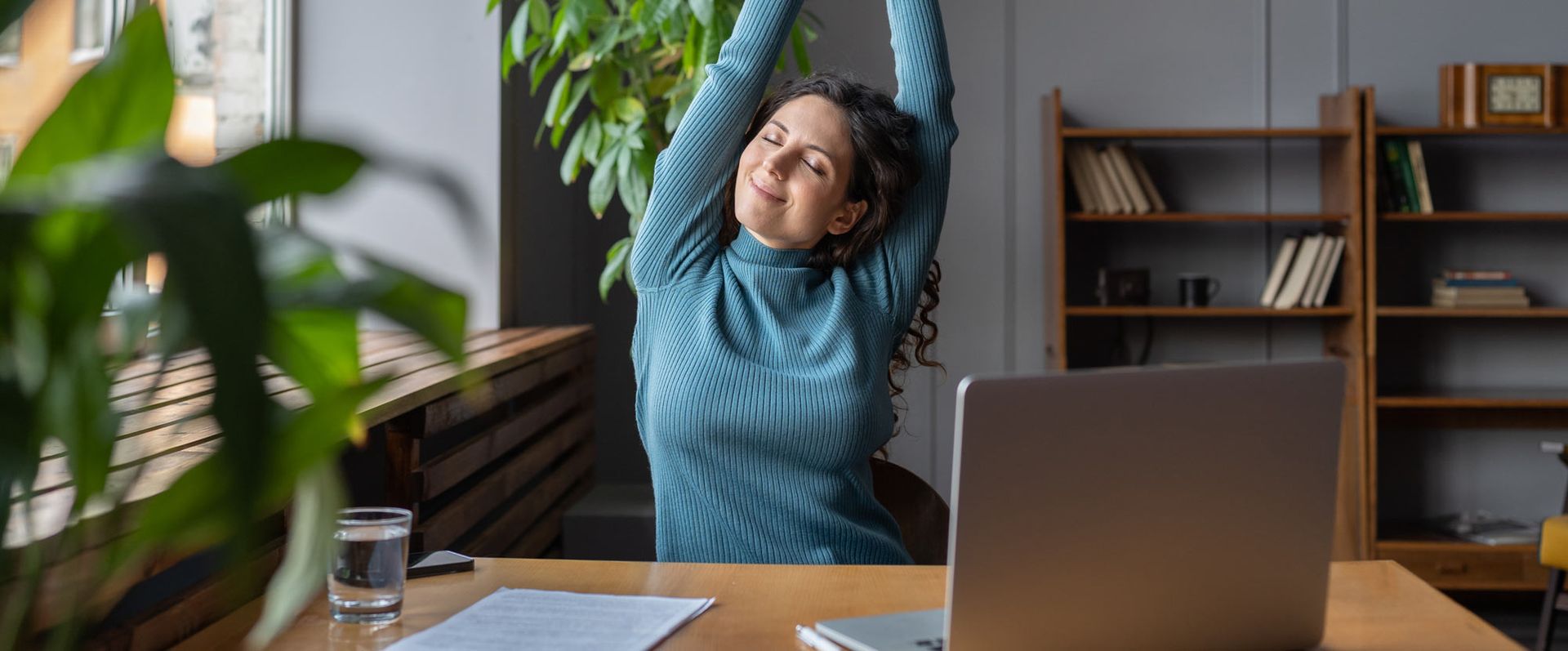 A woman is stretching her arms while sitting at a desk in front of a laptop computer. Introducing an Annual Holistic Physical