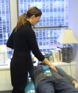 Dr. Alicia Armitstead doing nutrition response testing touches a person's chest. Indoors, by a window and desk with lamp.