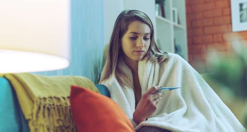Woman wrapped in blanket on couch, checking a thermometer, looking concerned. How To Avoid Getting Sick This Fall