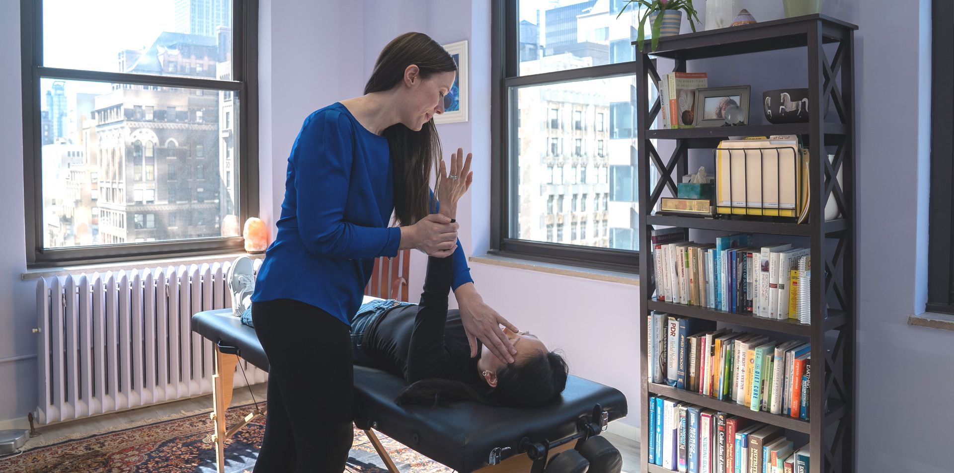 Dr. Alicia Armitstead in blue shirt stretches arm of person lying on massage table. Bookshelf and window in background explaining How Nutrition Response Testing Works