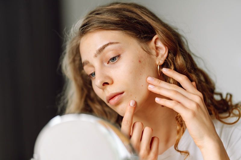A woman is inspecting her skin in a mirror, her hand resting on her cheek with concern.