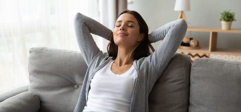 Woman relaxing on a couch with her arms behind her head, eyes closed, in a living room. The Science of Stress Relief 