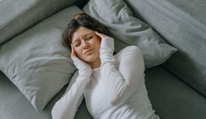 Woman lying on a gray couch, holding her temples, looking distressed. Why Root-Cause Care Matters for Chronic Conditions: Healing Beyond Symptoms