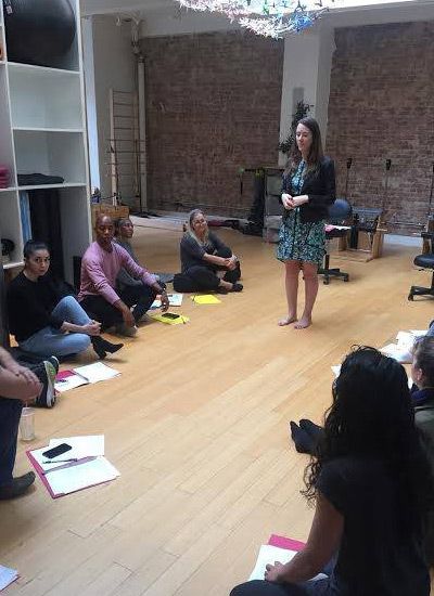 Dr Armitstead stands barefoot, addressing a group seated on the floor. Light wood floor, exposed brick wall.