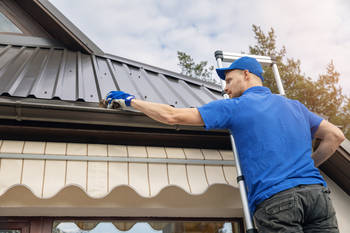 A man is standing on a ladder fixing a gutter on a roof.
