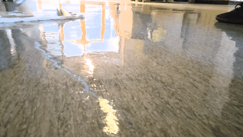 A low-angle view of a person using a squeegee to spread a glossy, liquid coating across a grey concrete floor.