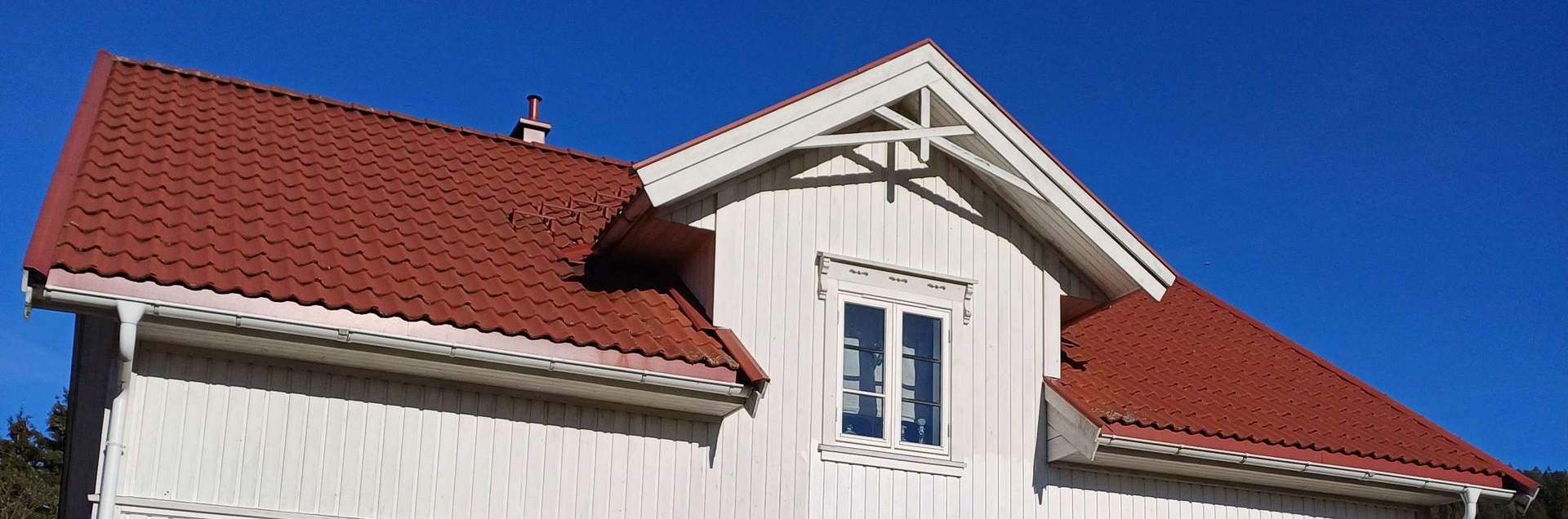 Et sidebilde av et hvitt trehus med rødt tegltak og et takvindu under en klar blå himmel.