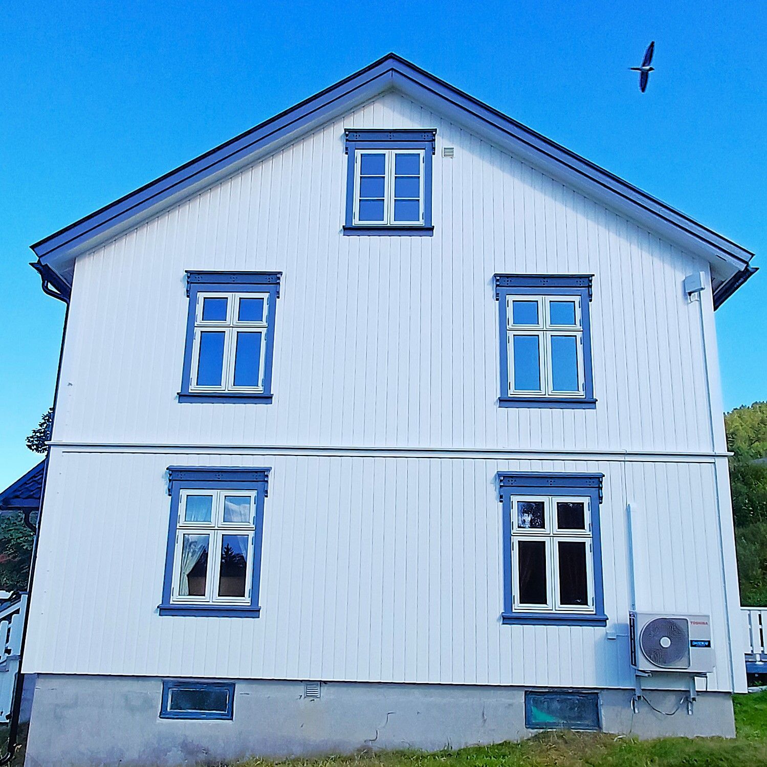 Et hvitt, toetasjes trehus med blå lister, fem vinduer, en grå steinfundament og en fugl som flyr på den blå himmelen.