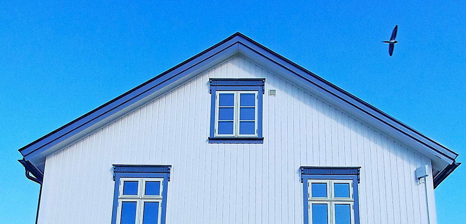 Et hvitt hus med blå lister og tre vinduer ligger under en klar blå himmel, med en fugl som flyr øverst til høyre.