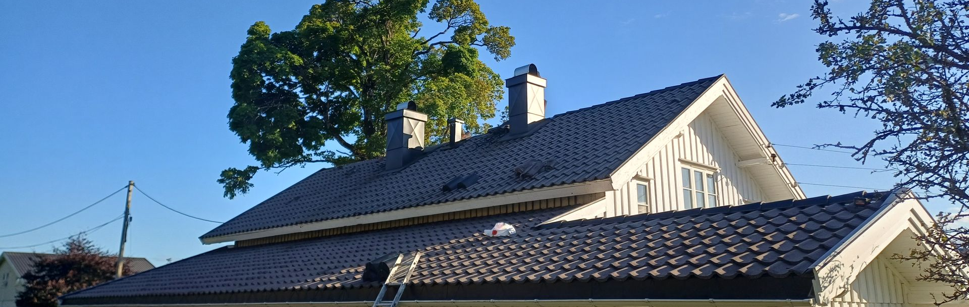 Et sidebilde av et hus med mørkt tegltak, to skorsteiner og en stige som lener seg mot den nedre takdelen.