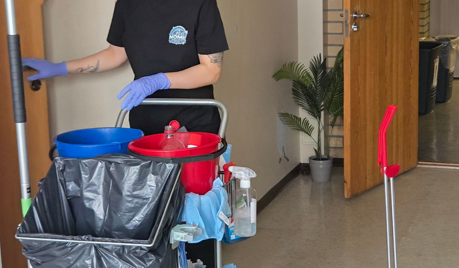 En person med blå hansker står med en rengjøringsvogn og rekker ut mot en lukket dør i et innendørs kontormiljø.