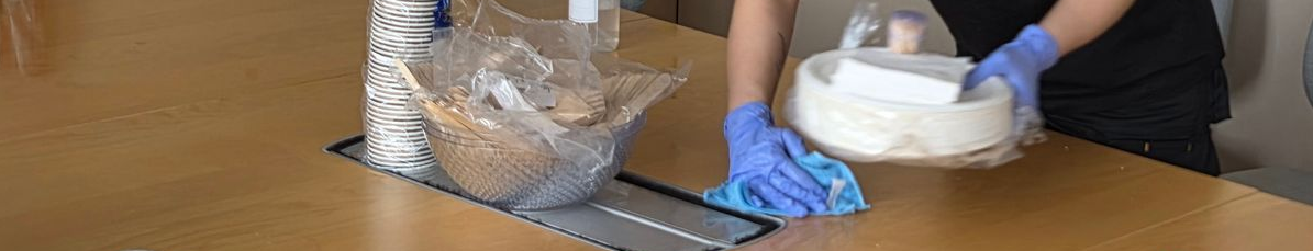 En person med blå hansker rengjør et konferansebord i tre, løfter en hvit bolle og flytter en blå rengjøringsklut.