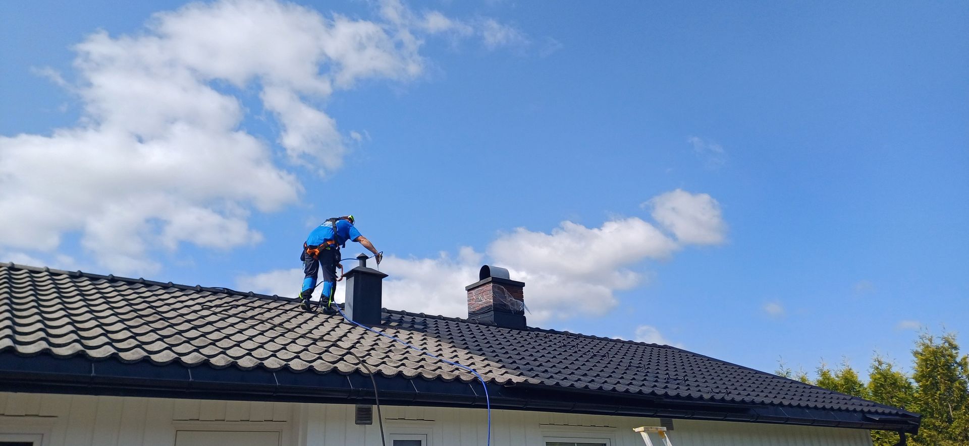 En person i blå arbeidsklær står på et mørkt tegltak og utfører vedlikehold på en skorstein under en blå himmel.
