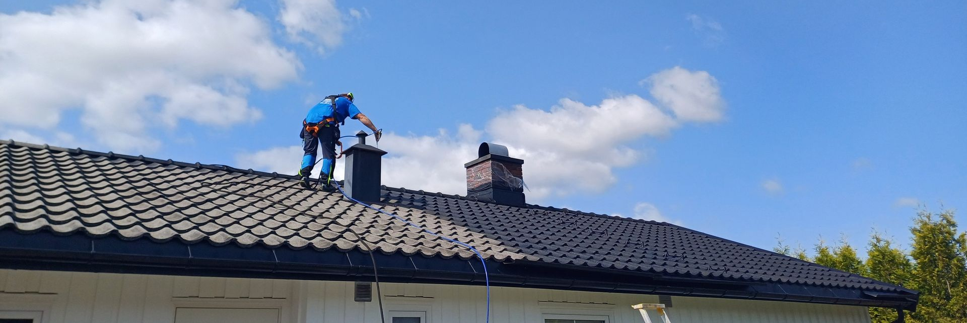 En arbeider i blå skjorte utfører vedlikehold på en skorstein oppå et mørkt tegltak under en lys, overskyet himmel.
