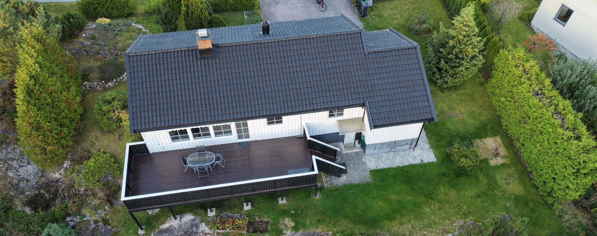 Luftfoto av et hvitt hus med mørkt saltak og en romslig mørk treterrasse i en frodig, grønn bolighage.