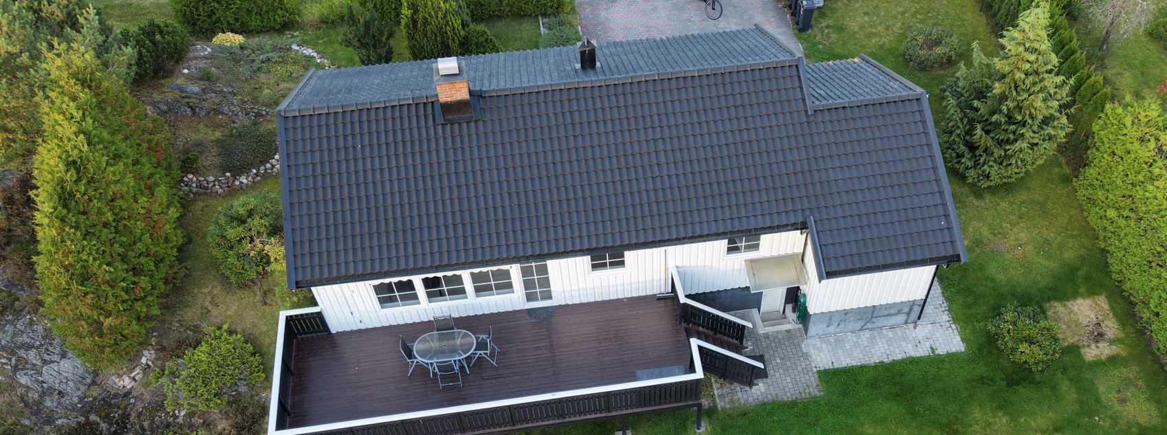 Luftfoto av et hvitt hus med svart tak og stor treterrasse i en grønn hage.