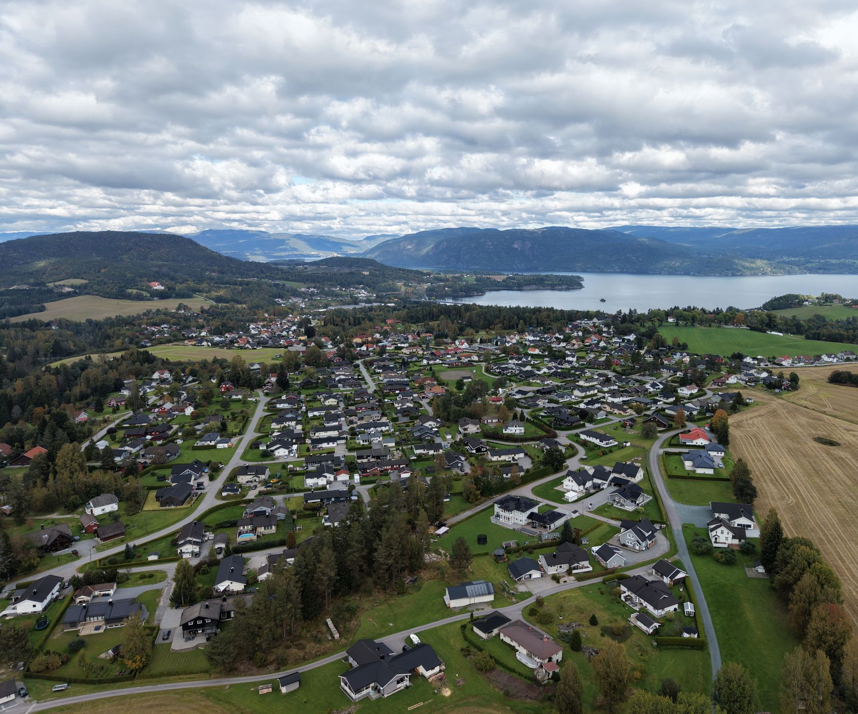 Luftfoto av et boligområde med hus og trær, beliggende nær en innsjø med fjerne fjell.