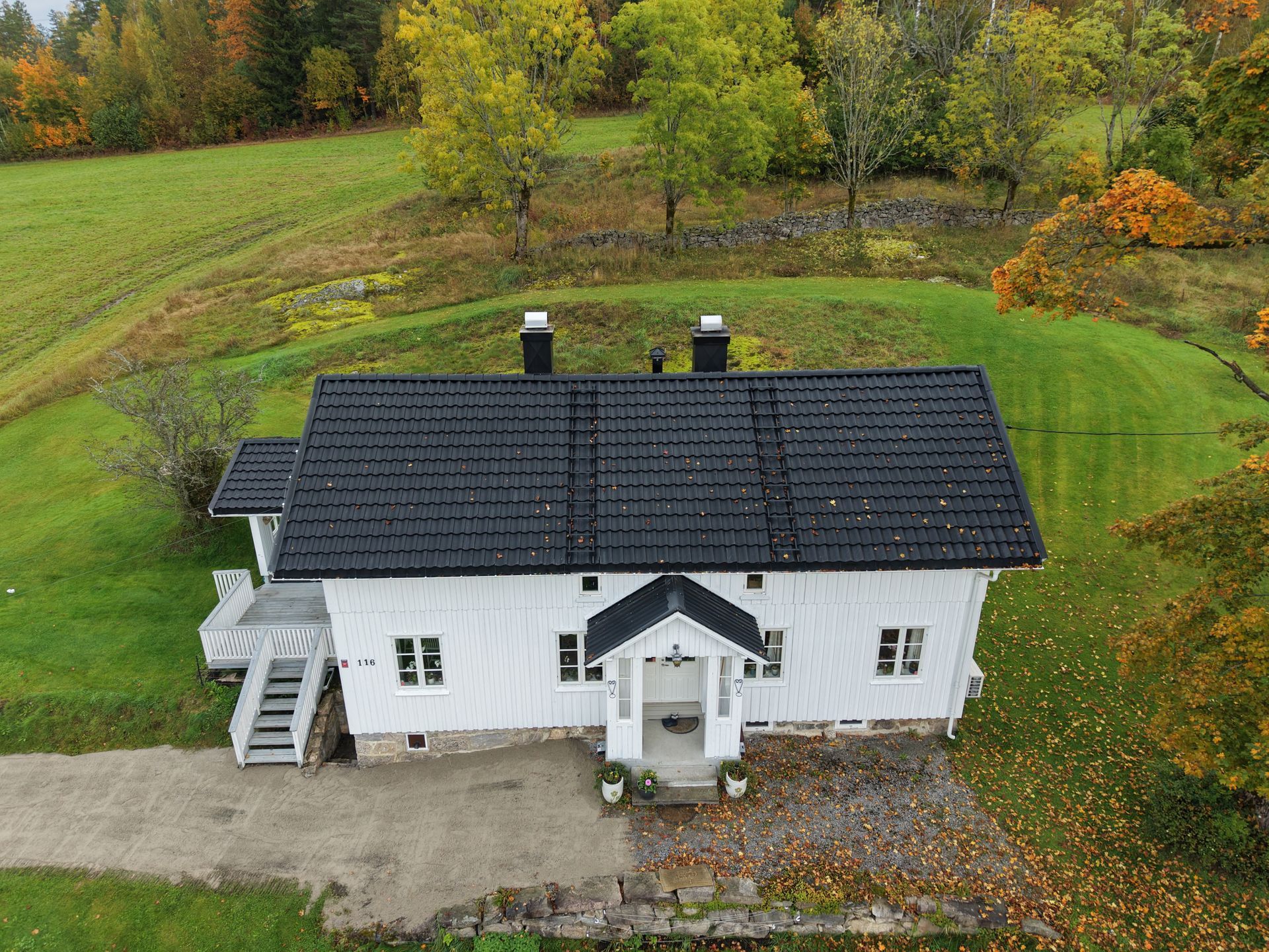 En luftfoto av et hvitt rektangulært hus med svart tak, veranda og liten trapp, beliggende i et gresskledd landskap.
