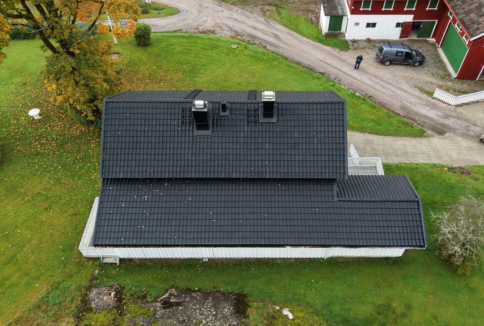 Luftfoto av et hus med svart tak og to skorsteiner, omgitt av en plen og en grusvei med en rød bygning i nærheten.