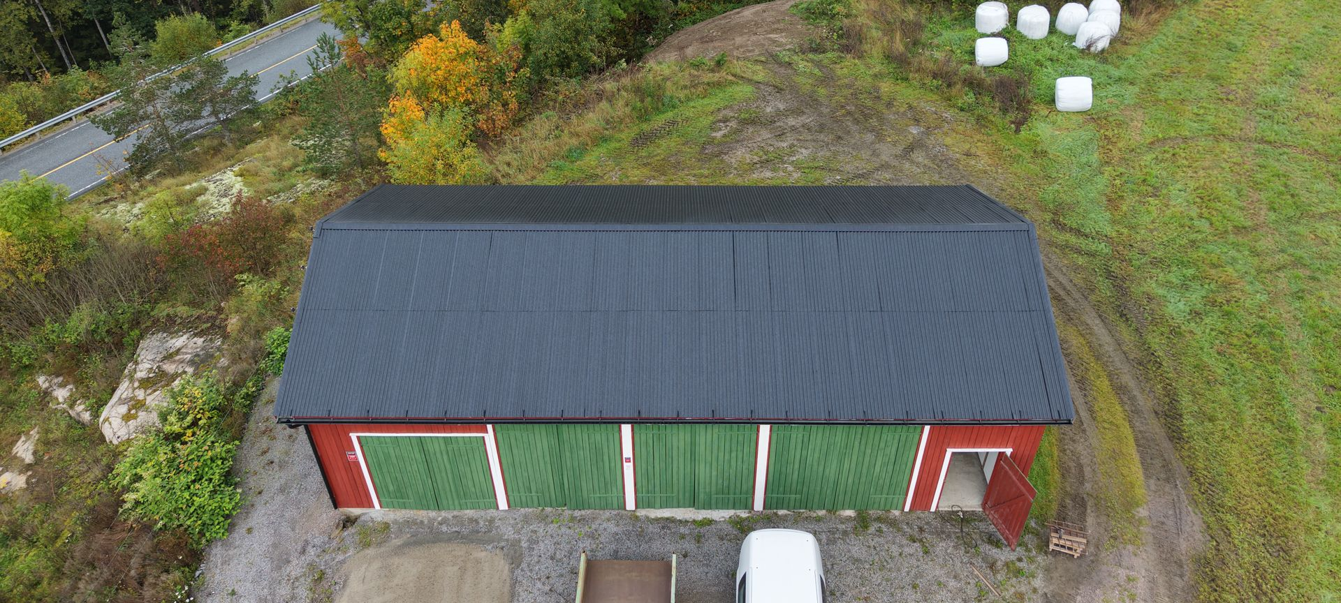 Luftfoto av en rød låve med svart metalltak, grønne dører og høyballer i et landlig landskap.