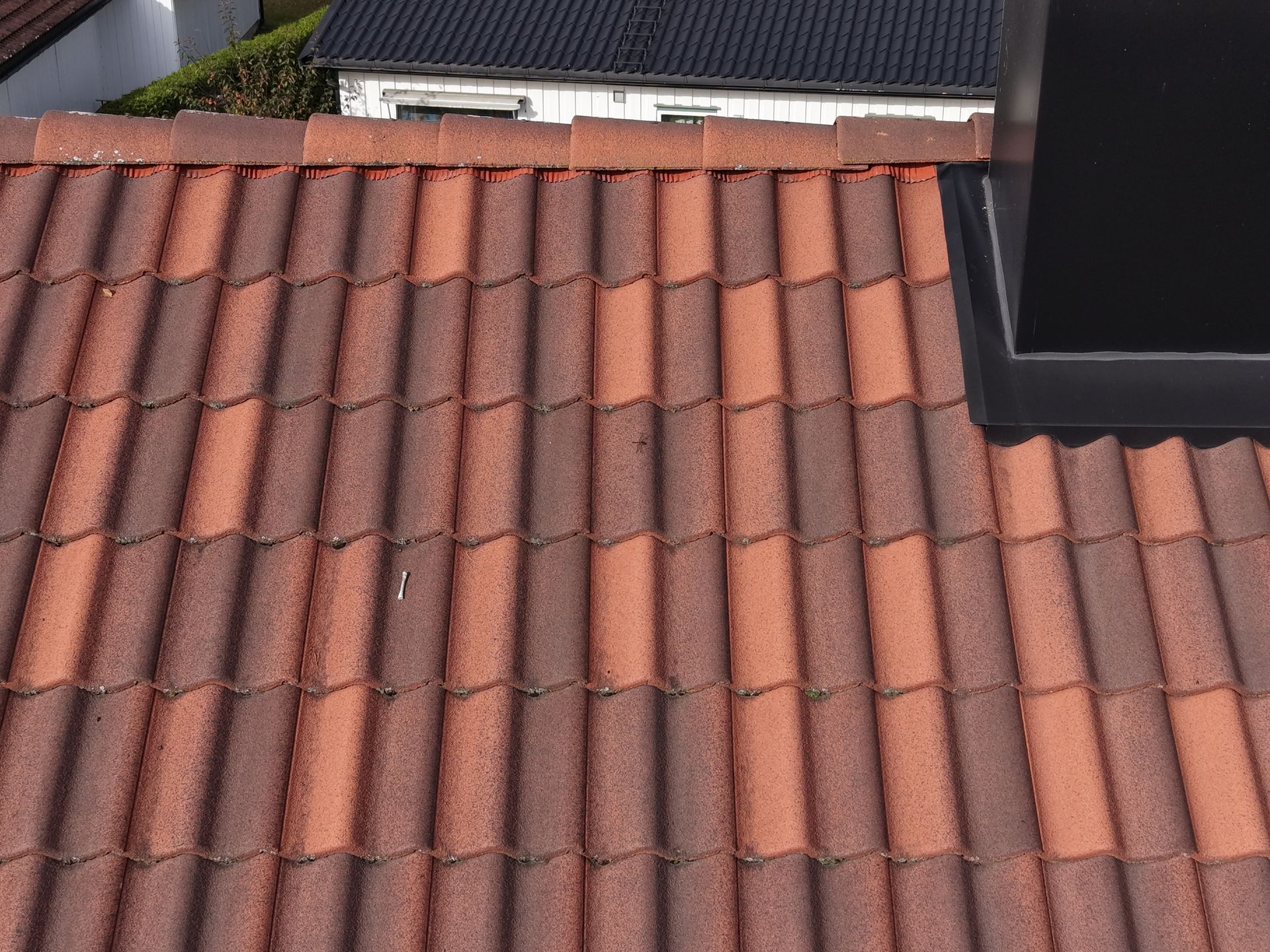 Et tak dekket av røde og brune, spraglete betongstein med en svart skorsteinsventil på høyre side.