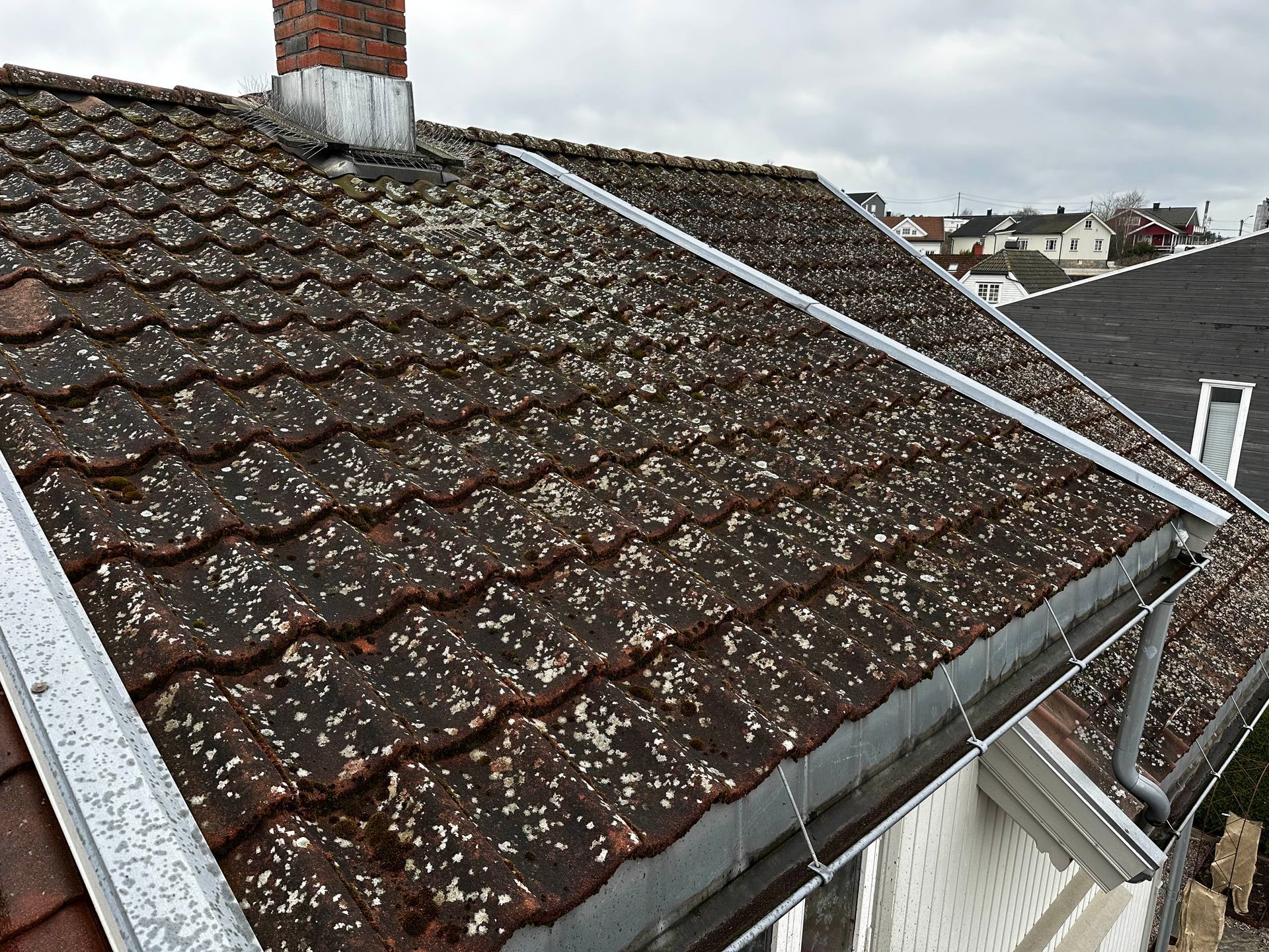 Et værbitt, mosedekket takstein med installerte metallrennebeskyttere og en skorstein mot en overskyet himmel.