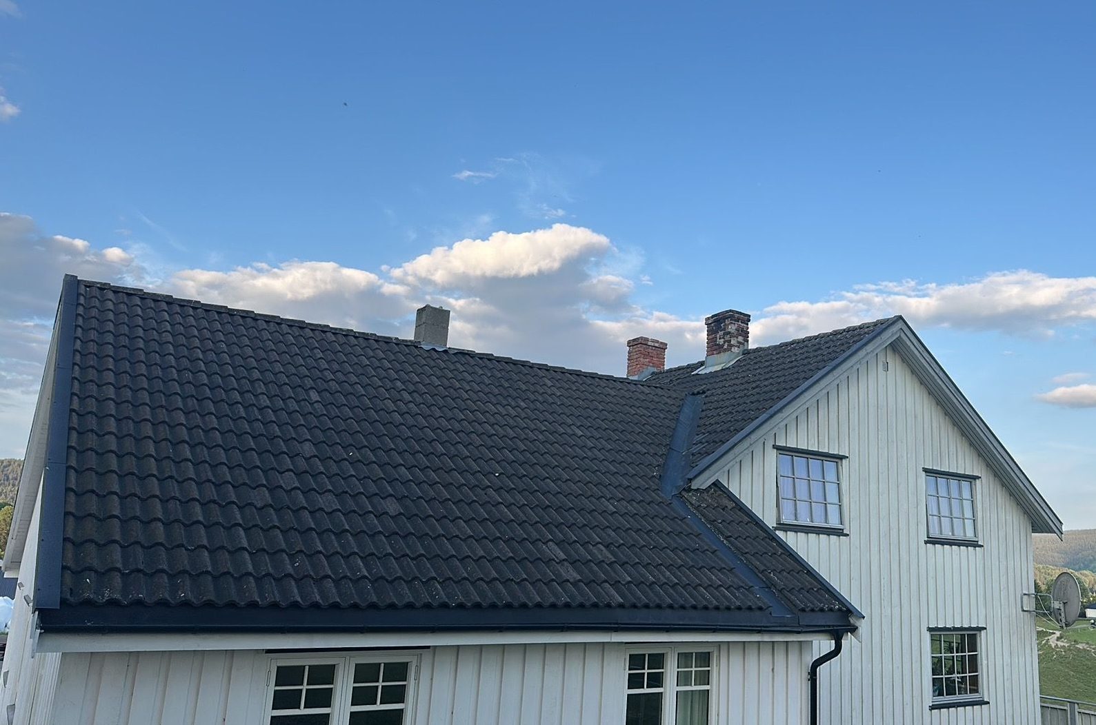Et hvitt hus med treverk og et svart tegltak med flere skorsteiner mot en knallblå himmel.