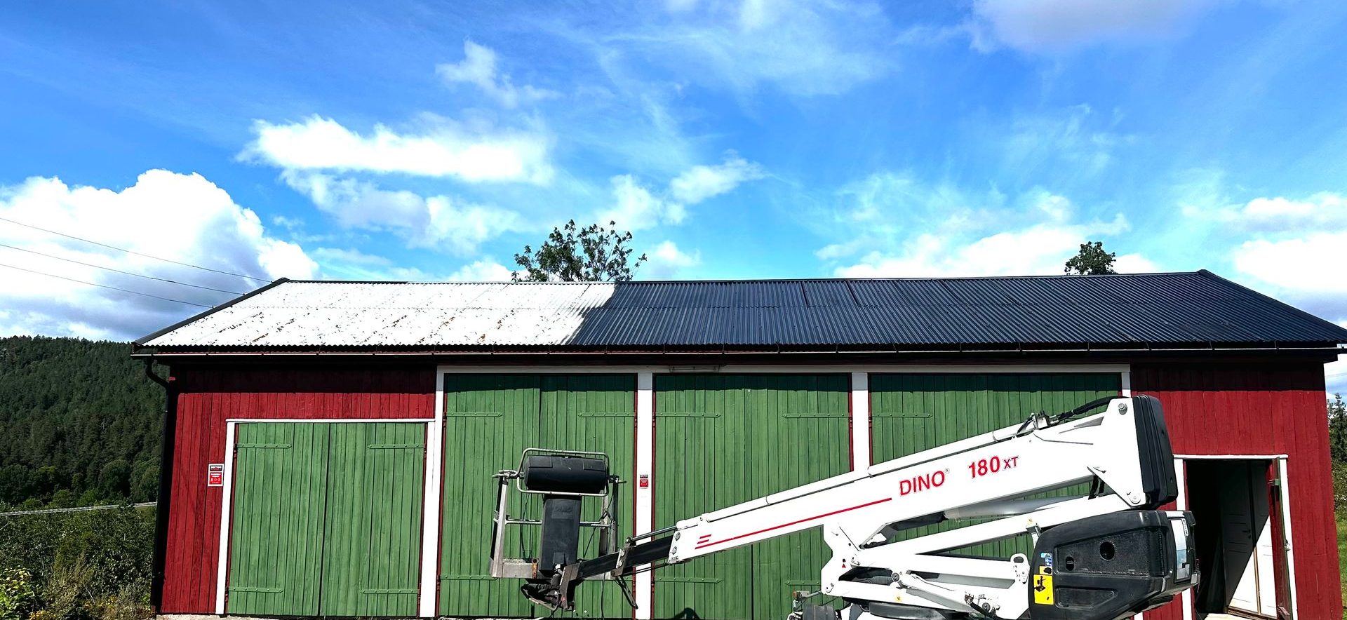 En rød bod med grønne dører under en blå himmel, med en hvit løfteplattform parkert foran under takrenovering.
