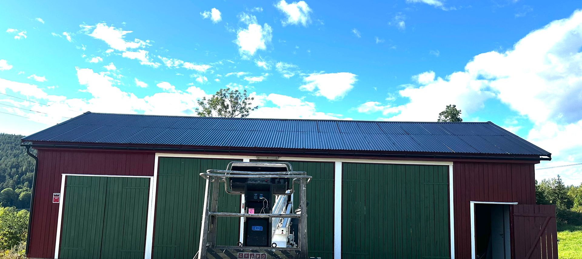En rød låve med grønne skyvedører og et svart tak, sett under en knallblå himmel med et maskineri foran.