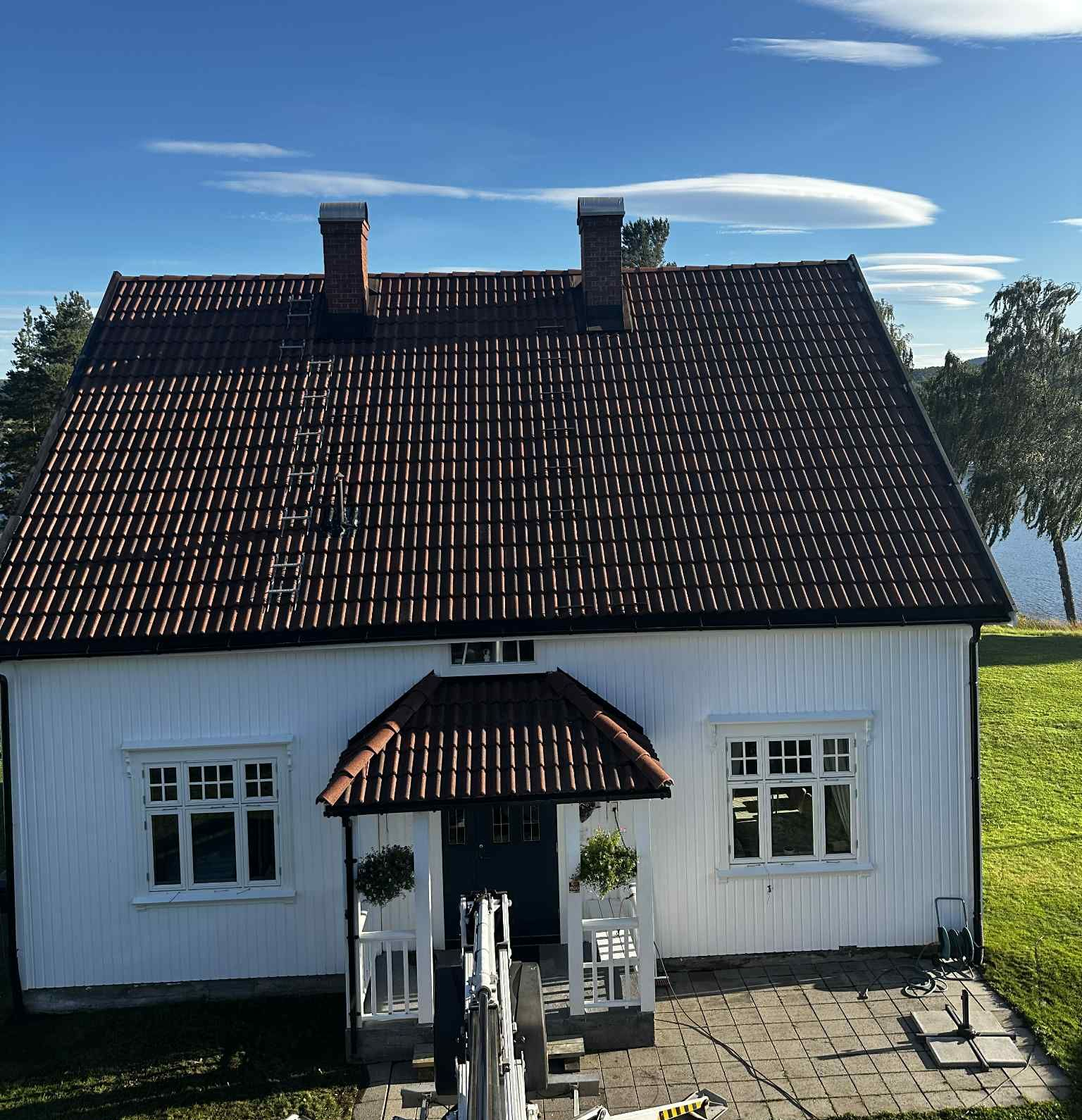 Et hvitt hus med mørkt tegltak, to skorsteiner og en liten veranda, mot en blå himmel med tynne skyer.