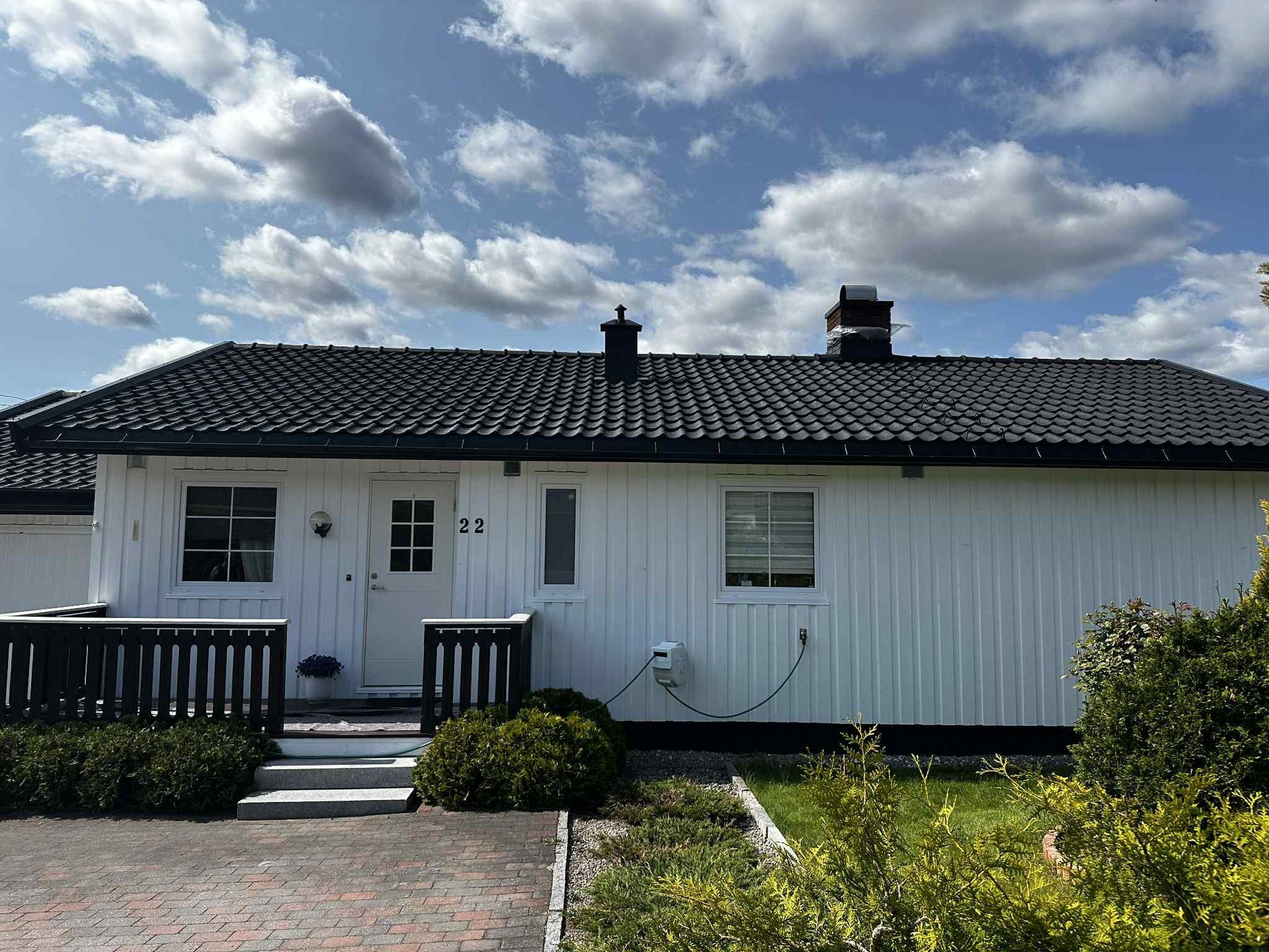 Et enetasjes hvitt hus med vertikal trekledning, svart tegltak og en liten terrasse foran under en overskyet blå himmel.