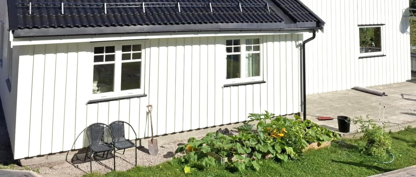 Et hvitt hus med vertikal kledning og svart tak, med en liten hagebed og to stoler på en grusterrasse.