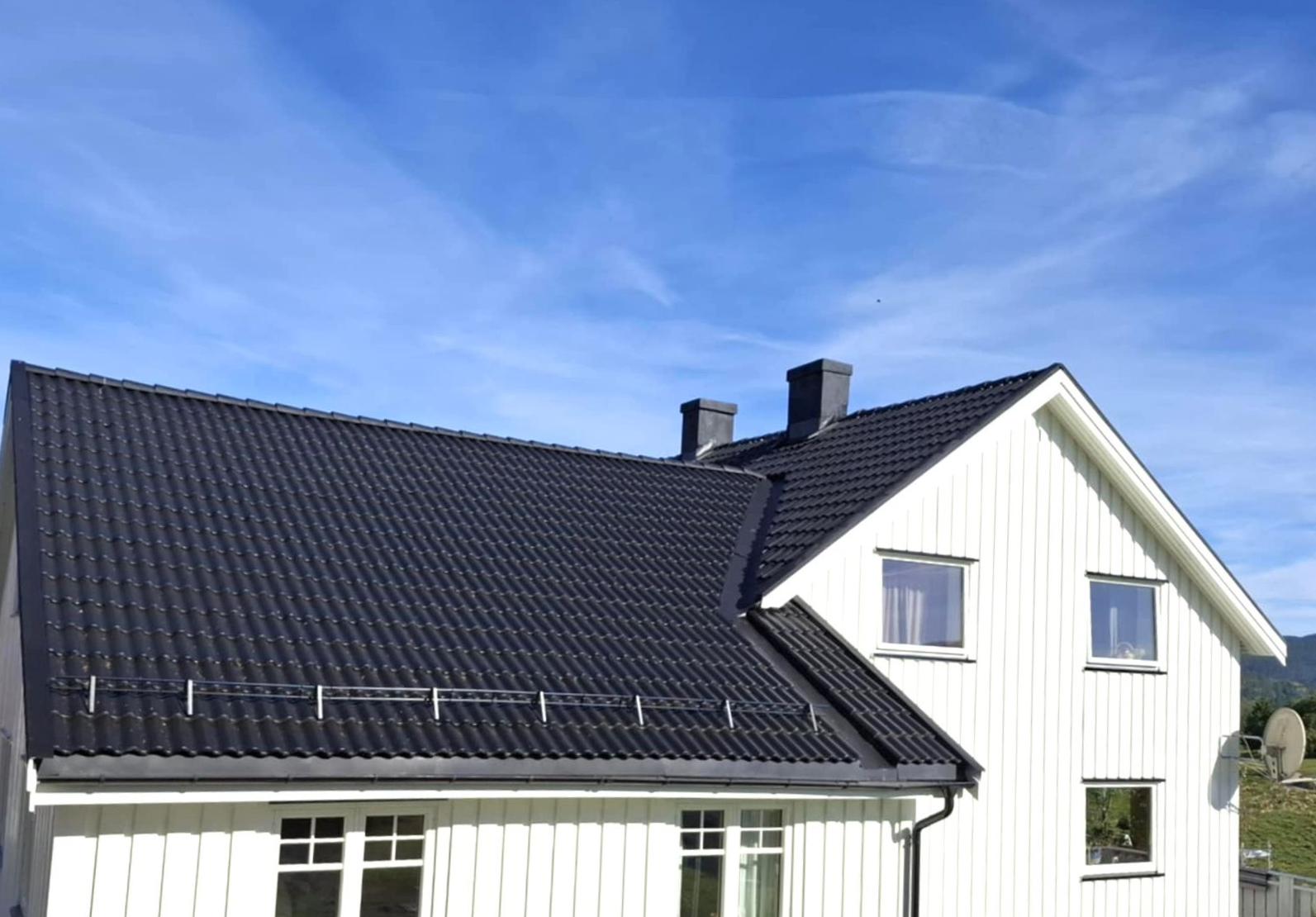 Hvitt hus med vertikal kledning og et mørkt, helvetestak under en klar blå himmel.