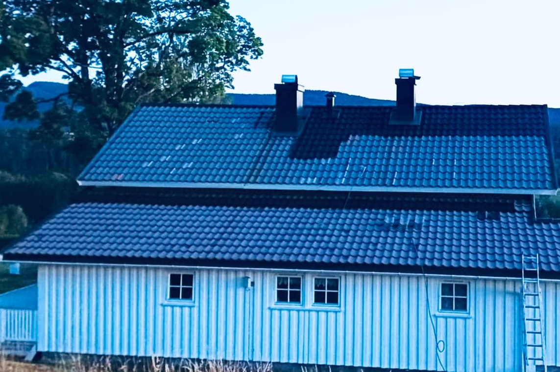 Et toetasjes hvitt hus med vertikal trekledning, et mørkt tegltak, to skorsteiner og en stige som lent seg mot veggen.